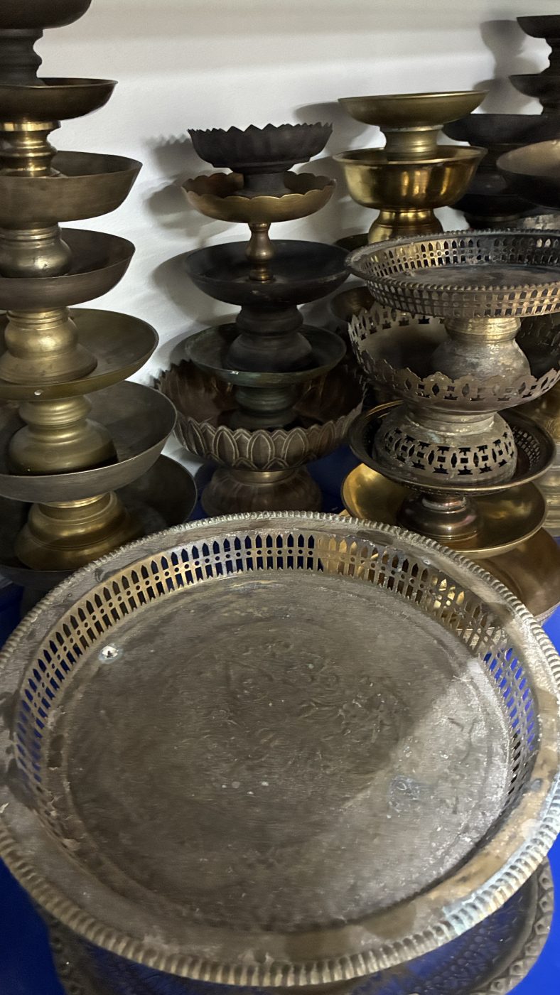 พานเชิงทองเหลืองหลายทรงเรียงซ้อนบนชั้นวาง มีถาดฉลุลายวางด้านหน้า